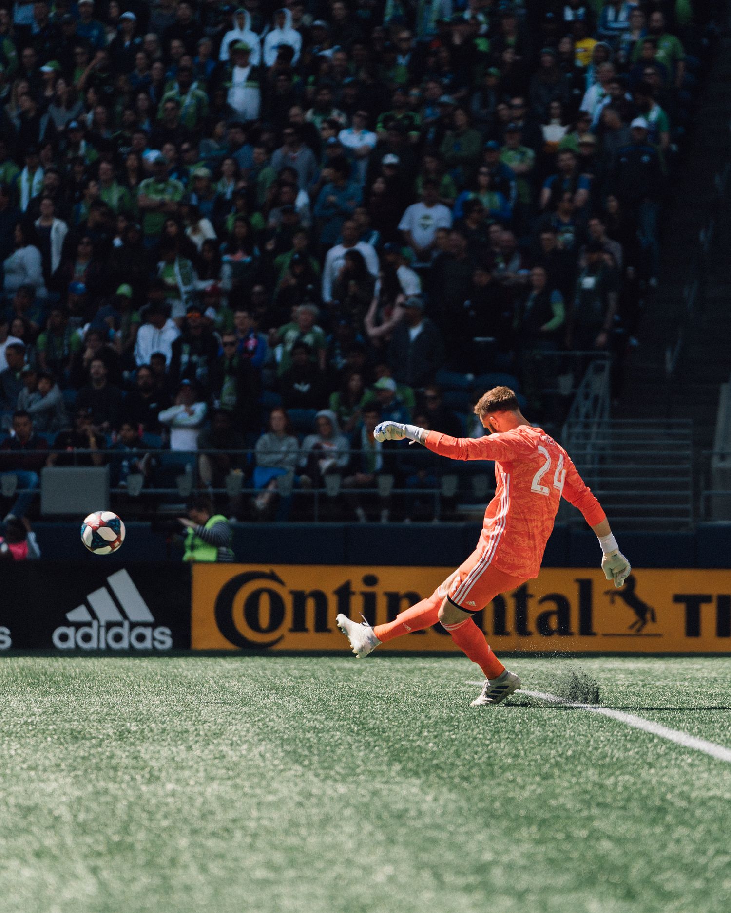 Soccer goalkeeper in an orange kit kicks the ball during a match as a packed stadium crowd watches behind adidas and Continental ad boards.