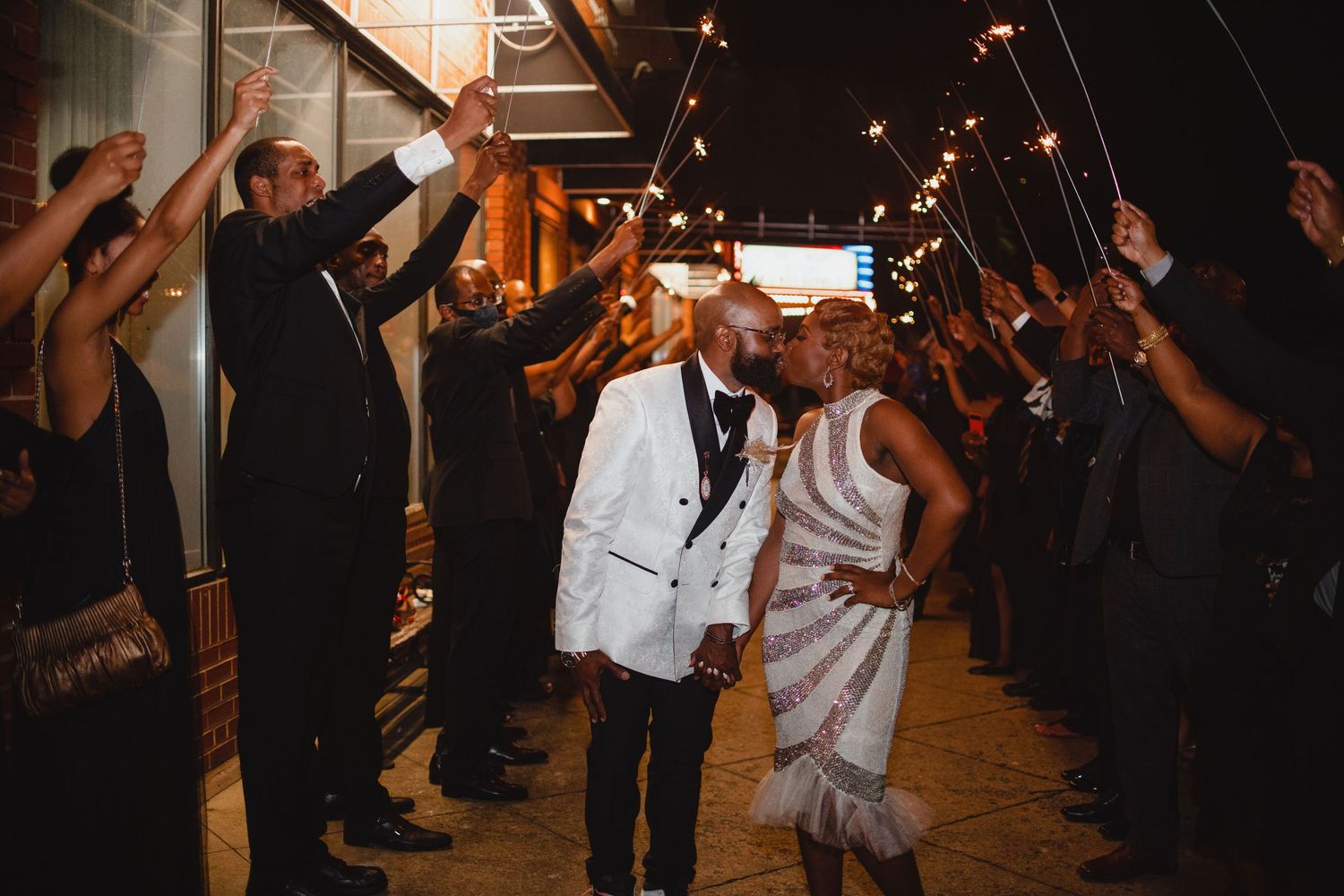 Newlyweds in a white tuxedo jacket and patterned dress walk through a sparkler send-off line of cheering guests at night.