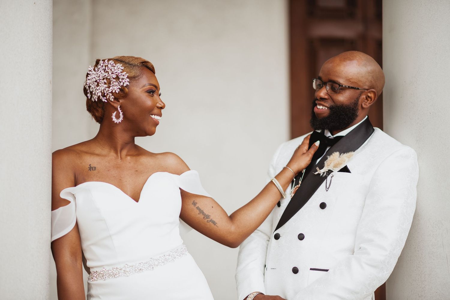 A smiling bride in a white off-the-shoulder gown with a floral hairpiece touches the lapel of her groom in a white tuxedo jacket.