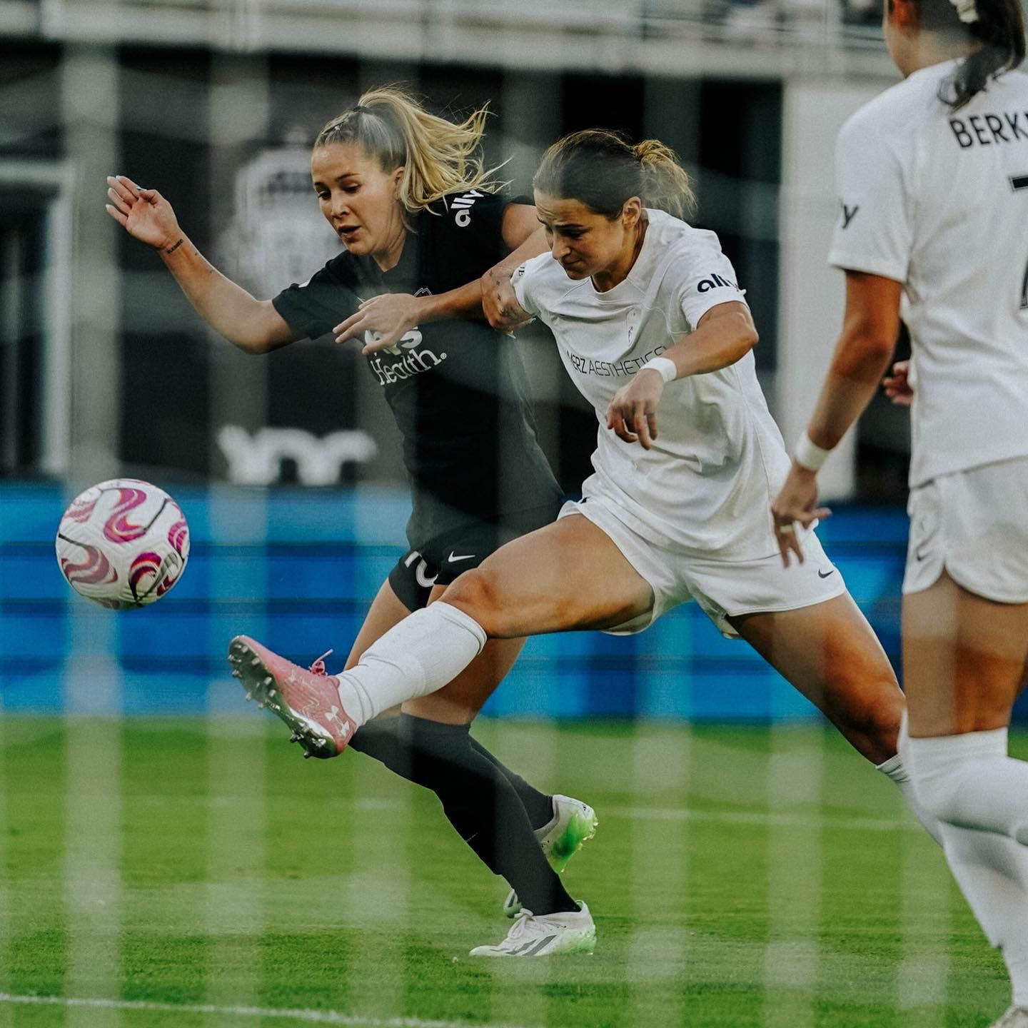 A player in a white kit kicks a soccer ball past a defender in black during a women's professional match.
