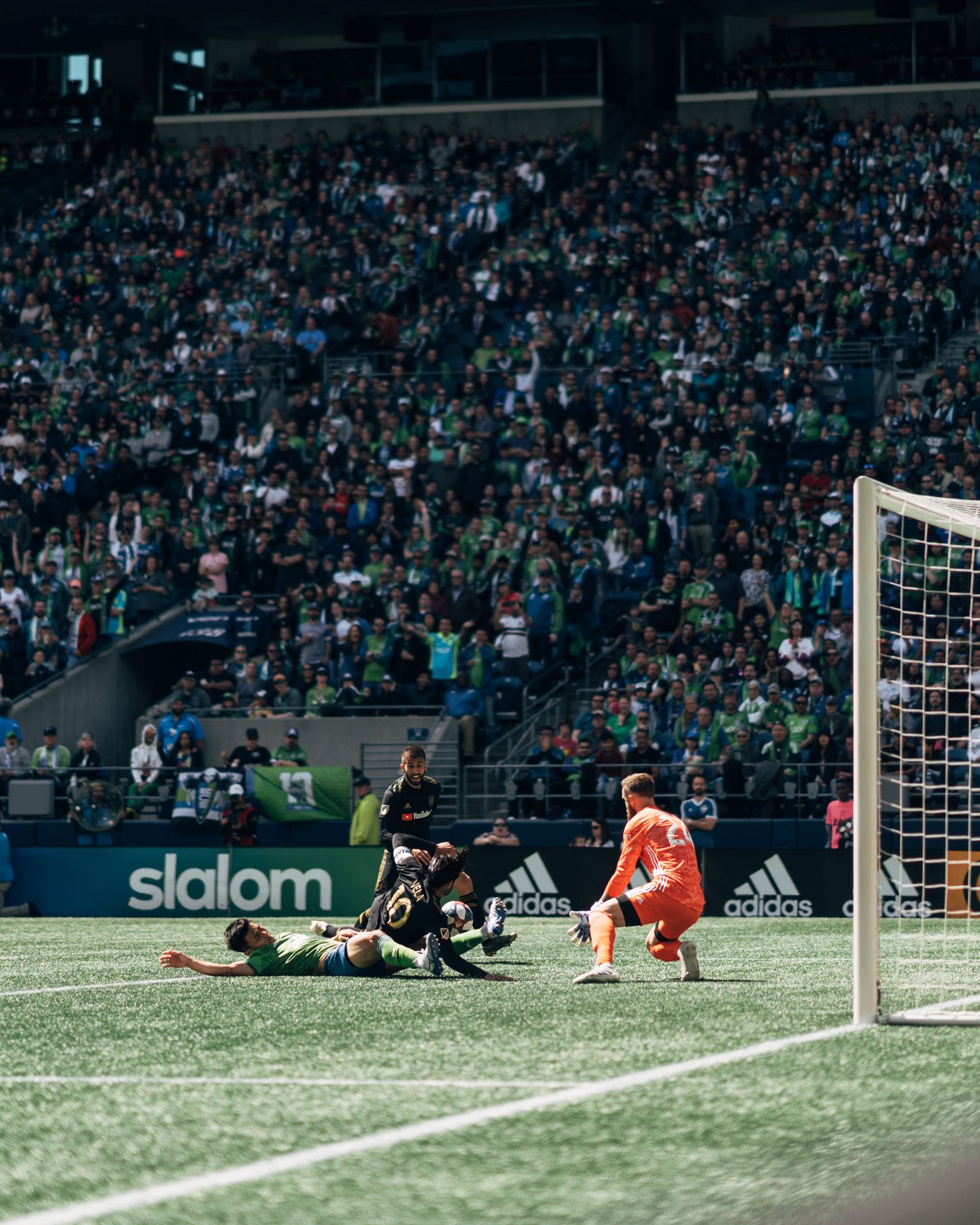 Soccer goalkeeper in orange stands near the goal as players collide on the grass before a packed stadium crowd in green.