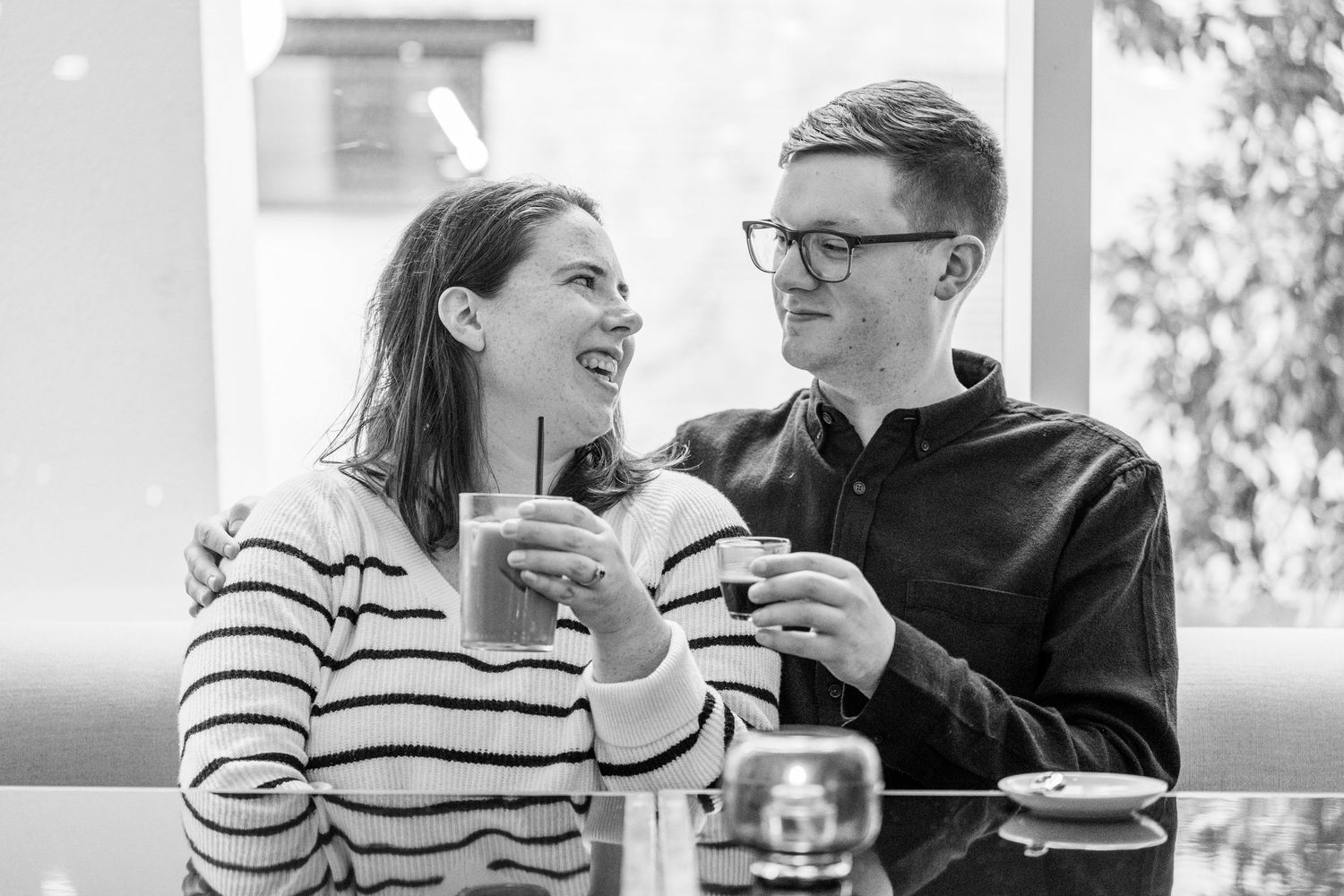 Black and white photo of a smiling couple sharing drinks and laughing together at a cafe table, the man wearing glasses.