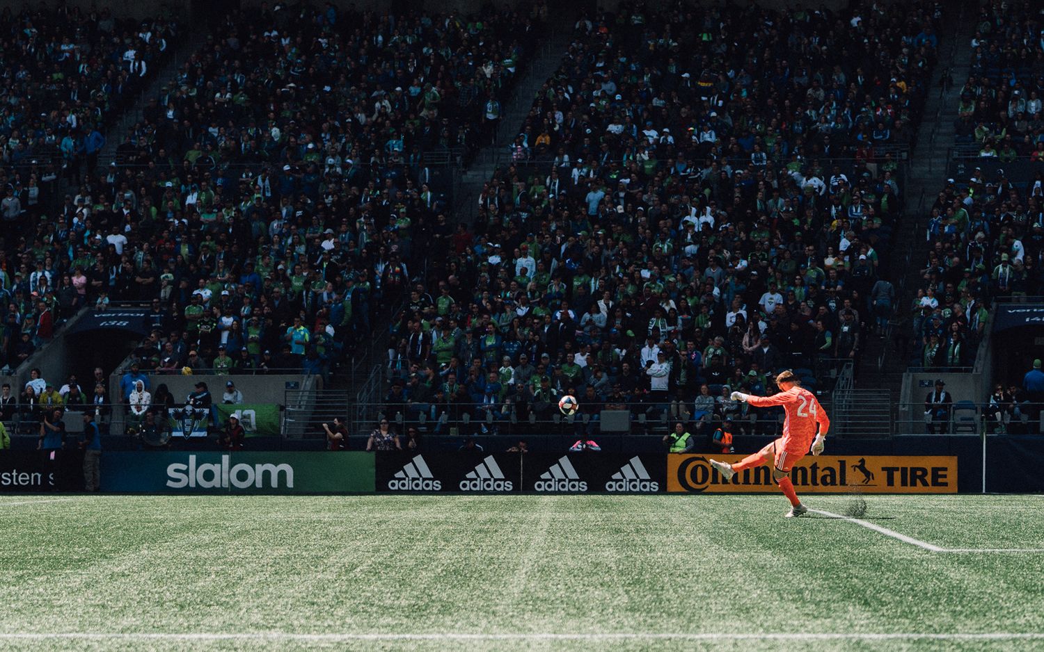 Goalkeeper in an orange kit takes a goal kick on a green pitch as fans fill the packed stadium stands behind him.