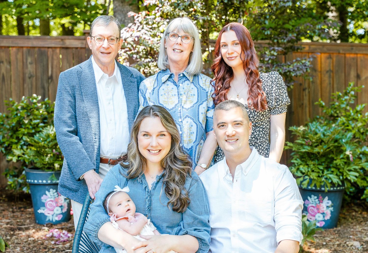 Multigenerational family of six, including grandparents and a baby, pose together in a lush green garden surrounded by flowering plants.