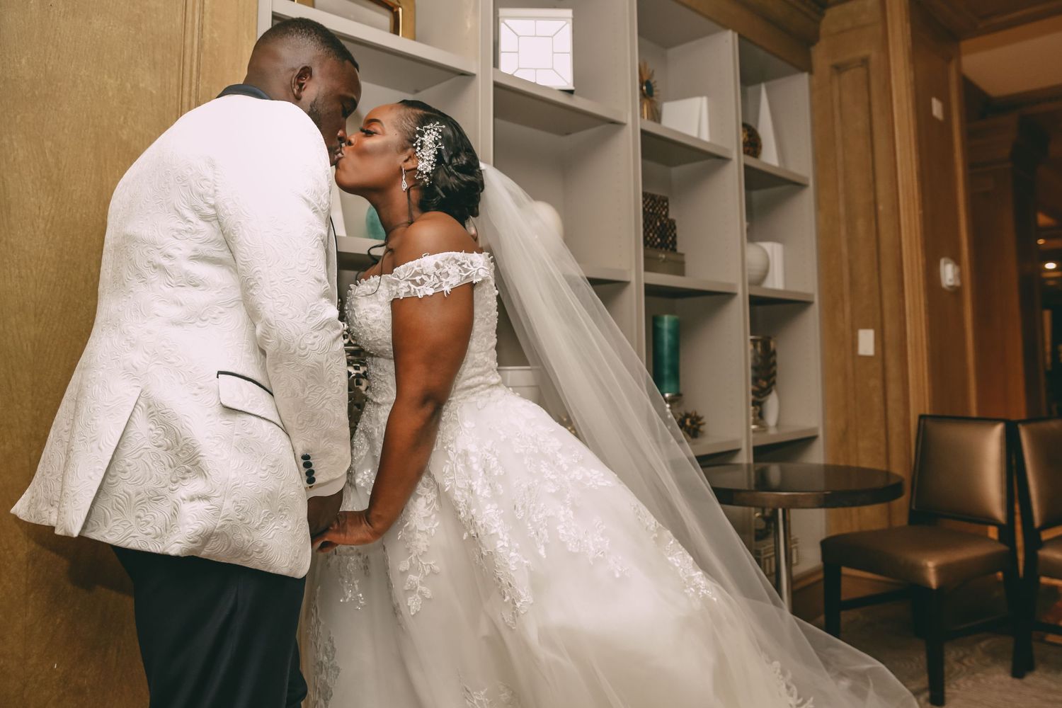 A bride in a lace-sleeved ball gown with a long veil kisses her groom in a white tuxedo jacket in an elegant room with built-in shelves.