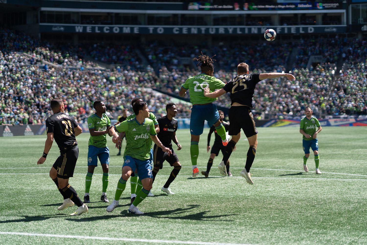 Soccer players in green and black kits leap for a header in front of the goal as the ball flies overhead in a packed outdoor stadium.