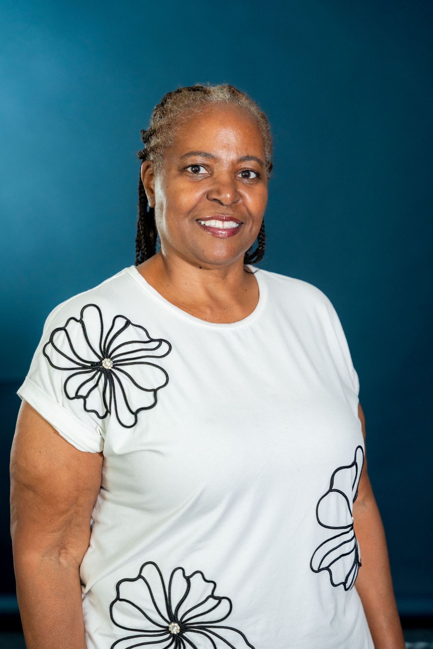 Smiling woman with braided hair wears a white top with black floral designs, posed against a deep teal background.