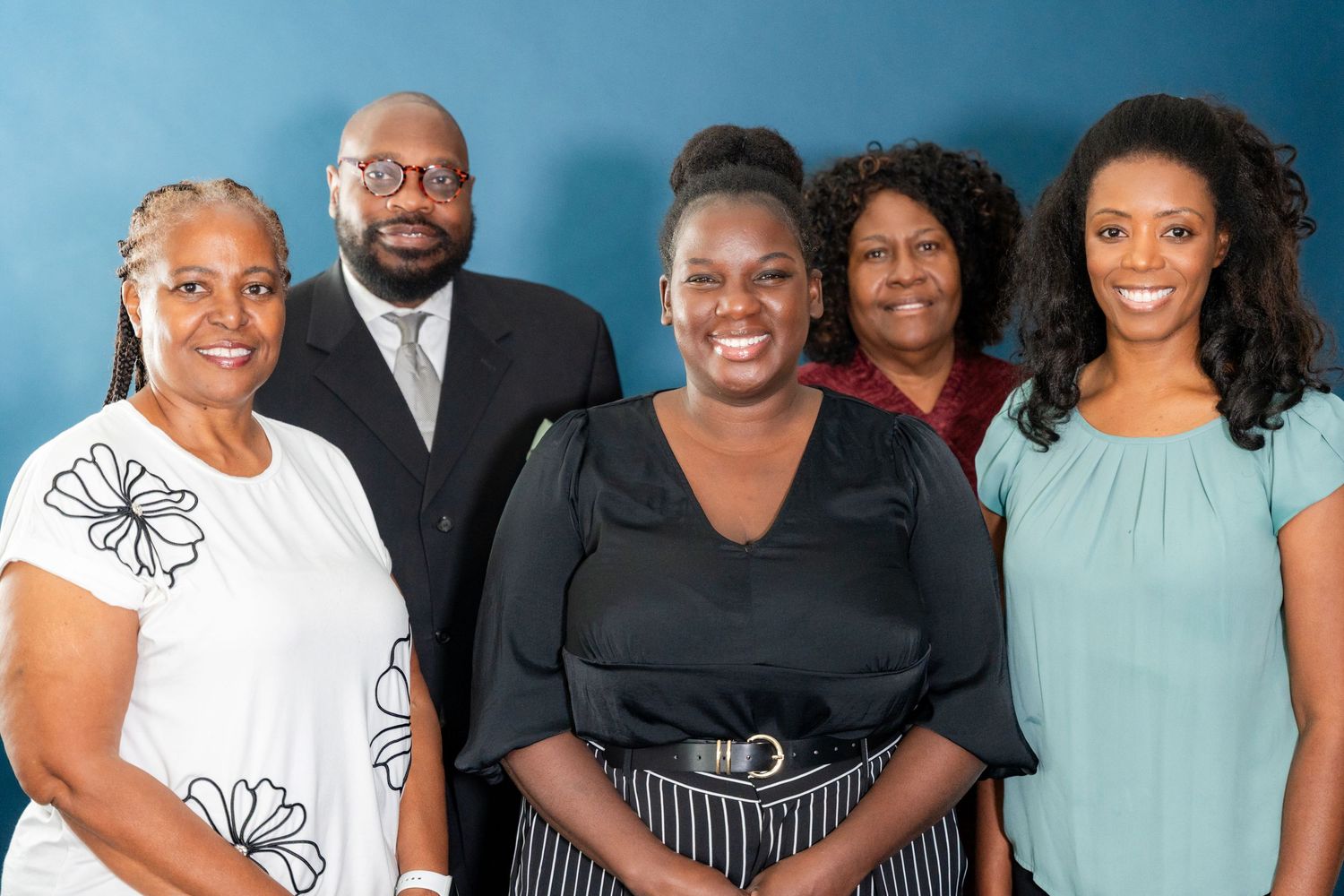 Five smiling colleagues, three women and a bearded man in a suit and glasses, pose together in a group portrait against a blue wall.