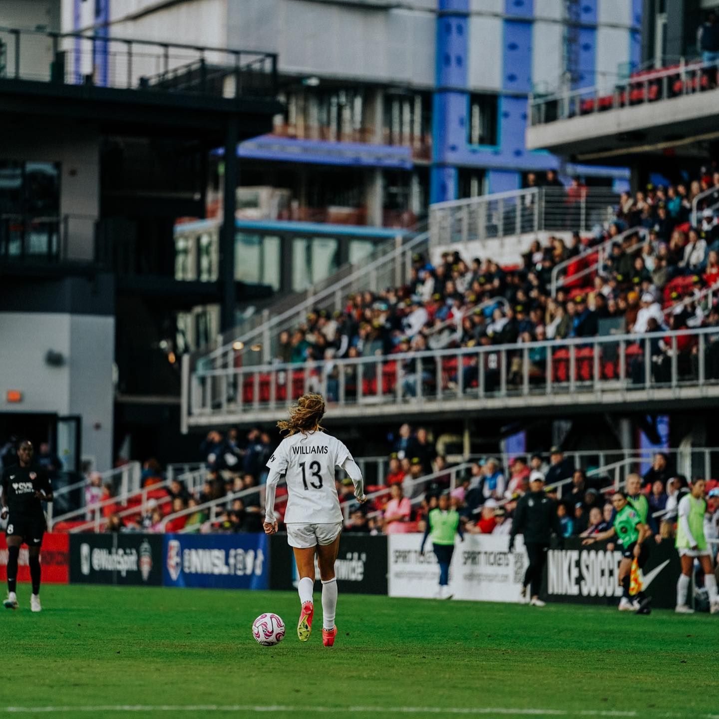 A soccer player wearing number 13 in a white kit dribbles the ball across the field as spectators fill the stadium stands.