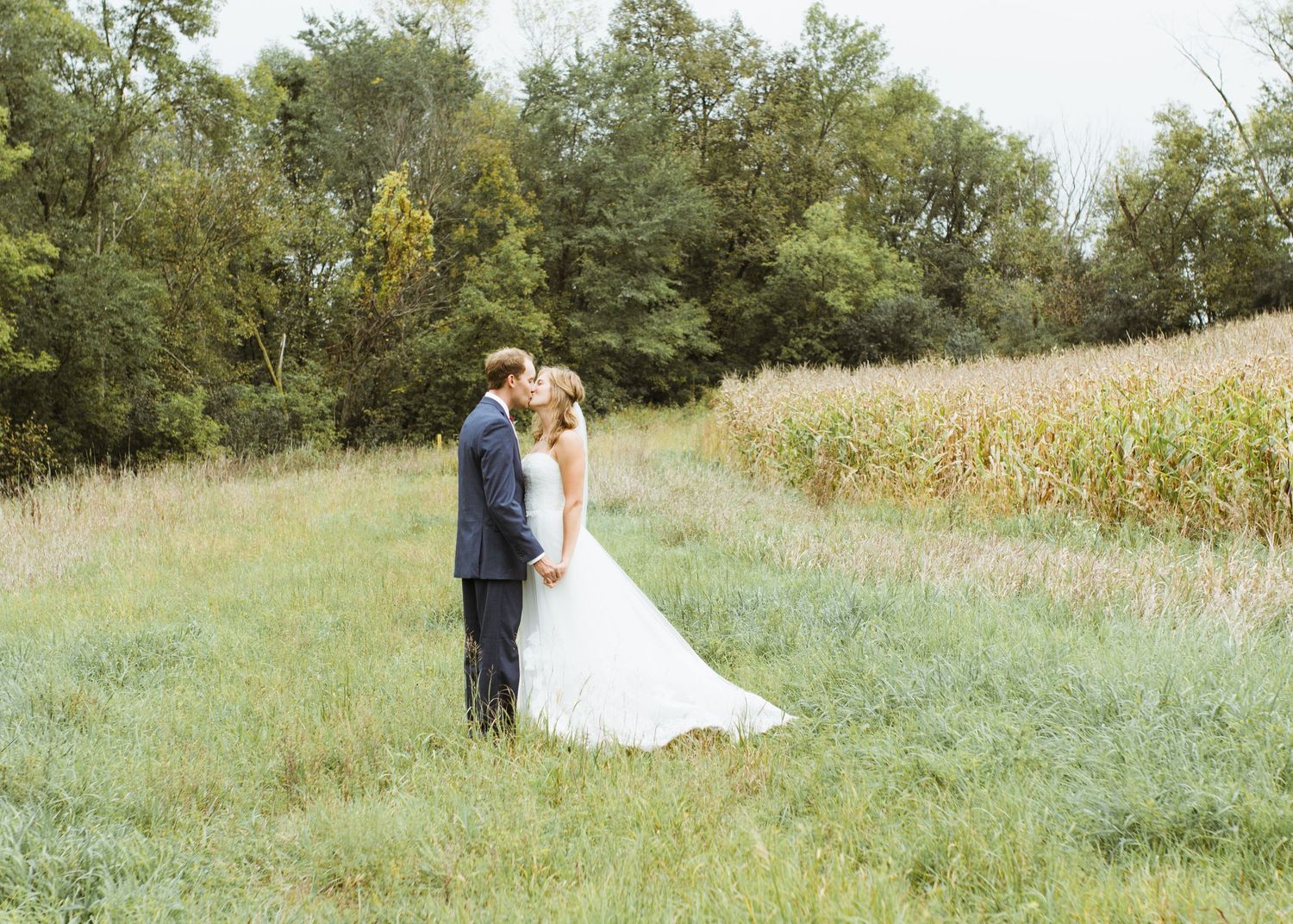 Bride and groom sharing a kiss in a lush green field with cornfields and trees in background.
