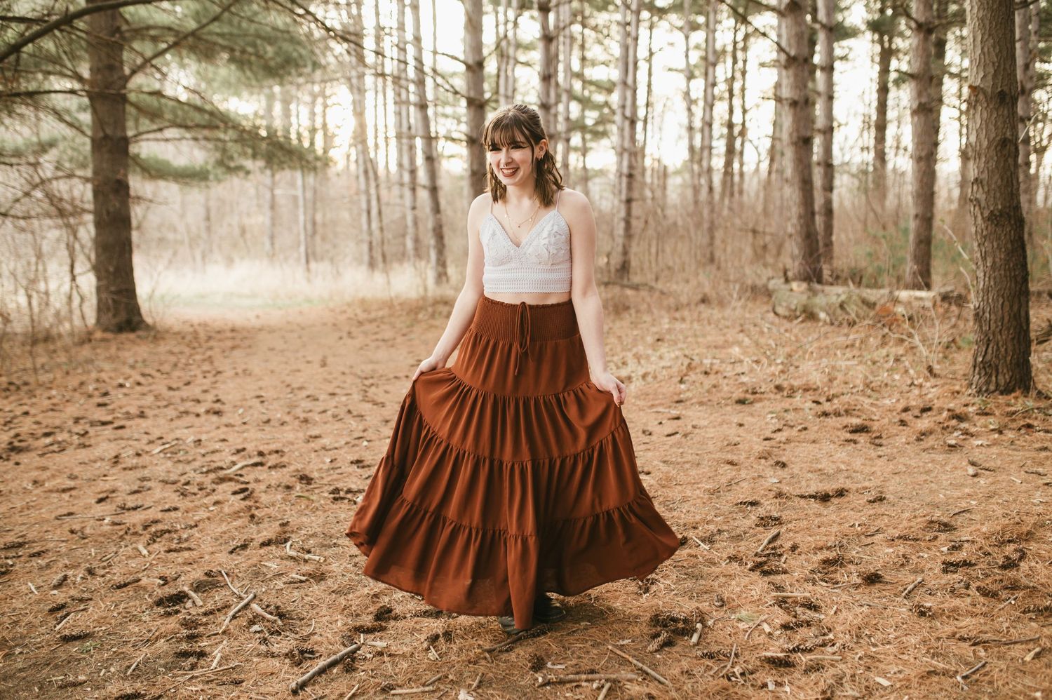 Young woman in rust tiered maxi skirt and white lace top walks through sunlit pine forest path.