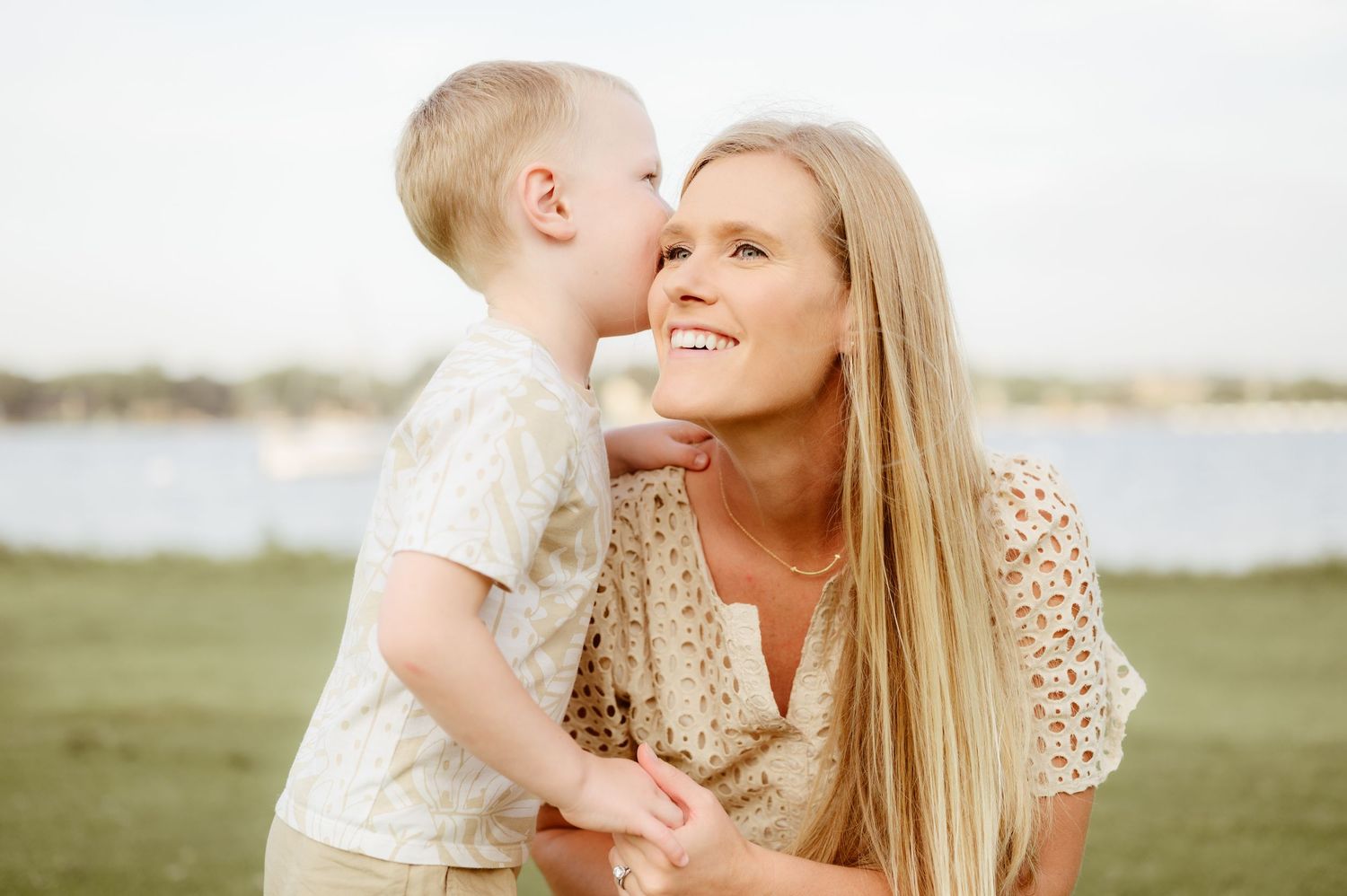 A blonde mother smiles as her young toddler gives her a kiss on the cheek outdoors.