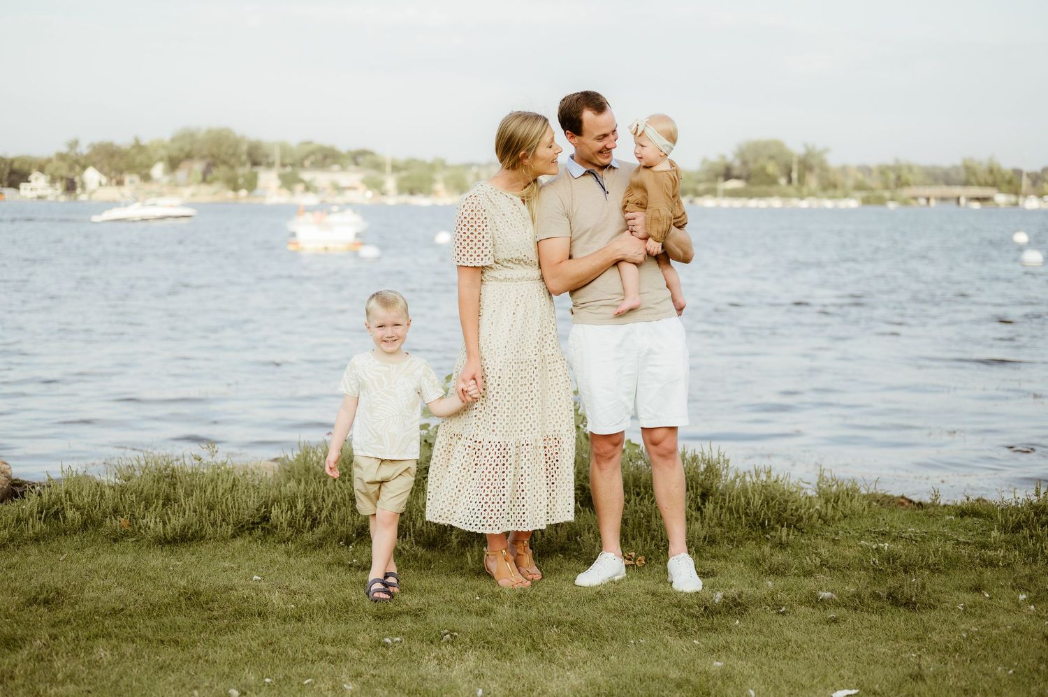 Happy family of four posing by lake minnetonka on a sunny day, parents holding toddlers on green grass.