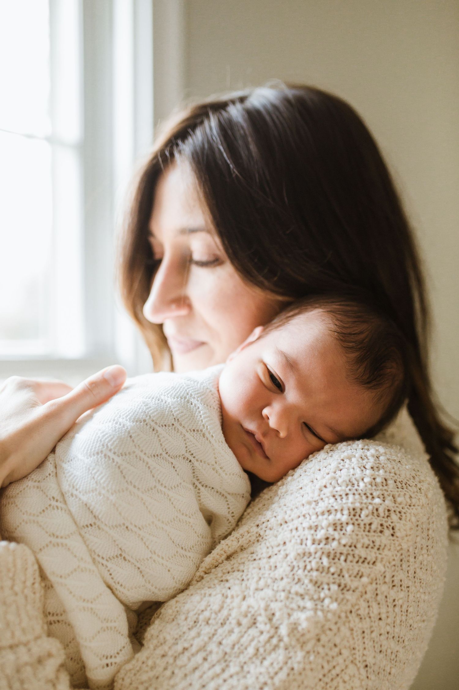 Dark-haired mother in cream knit sweater tenderly holding sleeping newborn baby against her chest by window.