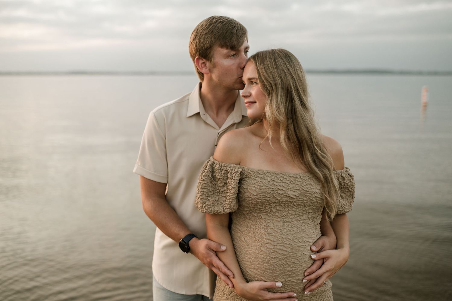 Loving couple embracing during a maternity photoshoot on a calm beach at golden hour.