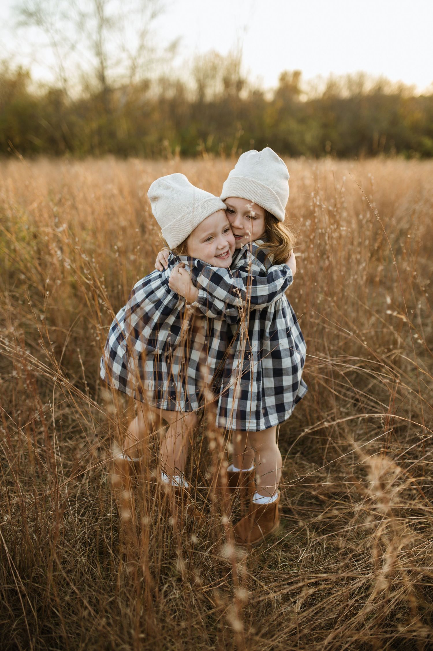 Two young siblings in matching plaid dresses and white beanies hugging in a golden autumn field.