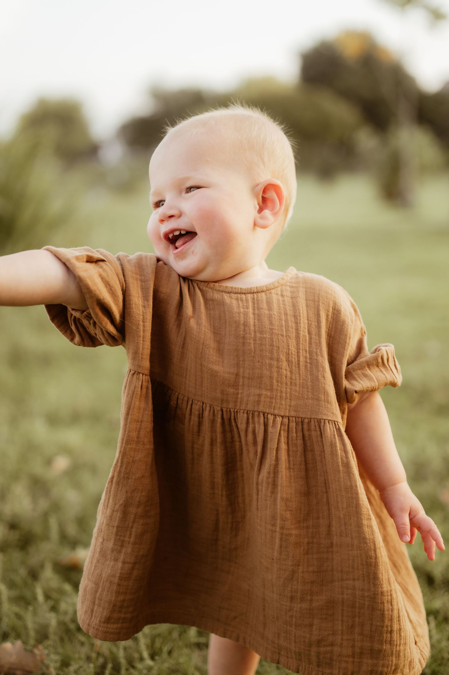 A joyful baby girl in a brown dress laughs while playing outdoors in a grassy field.