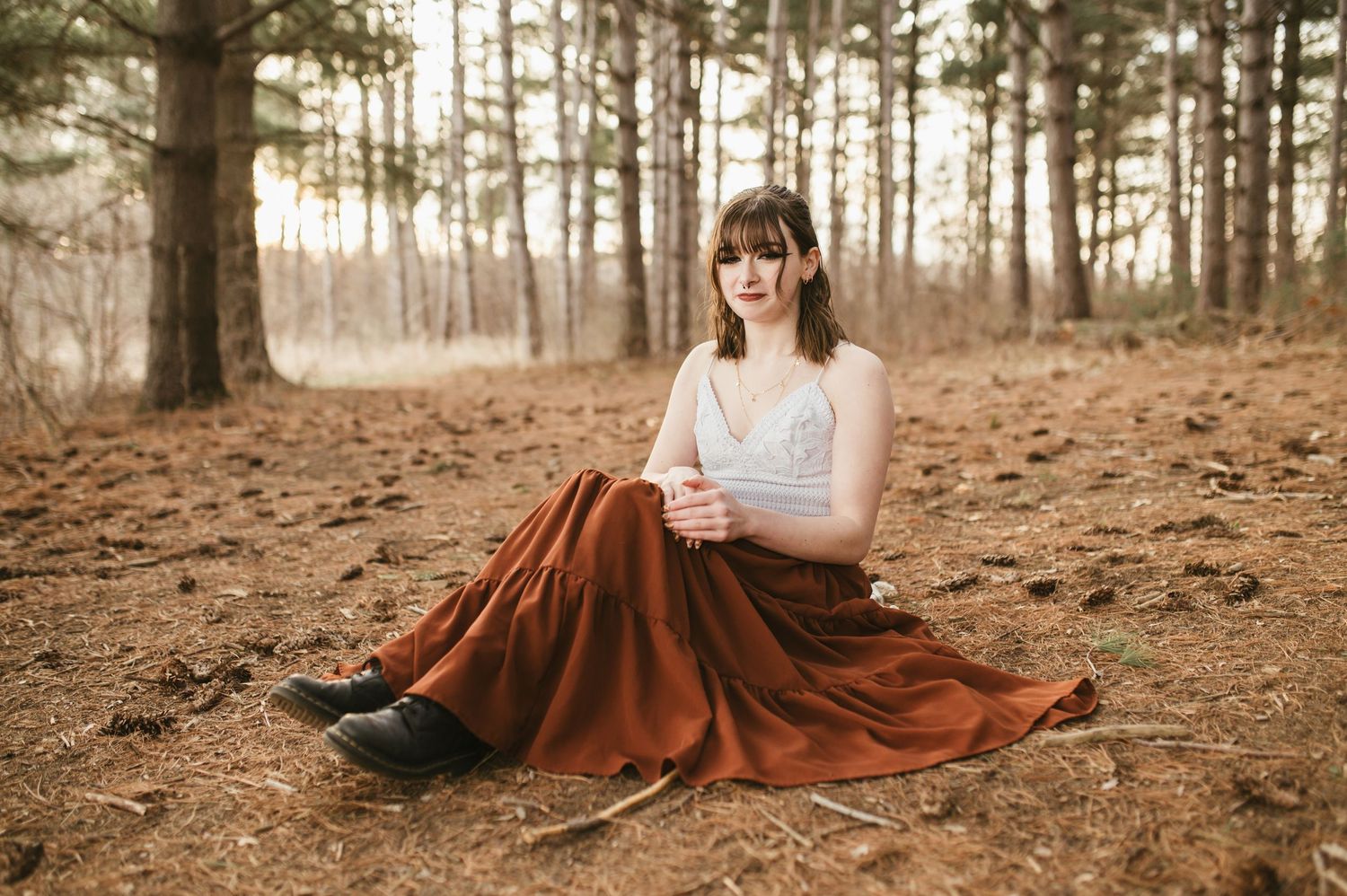 Young woman in rust maxi skirt and white lace top sits serenely on pine forest floor.