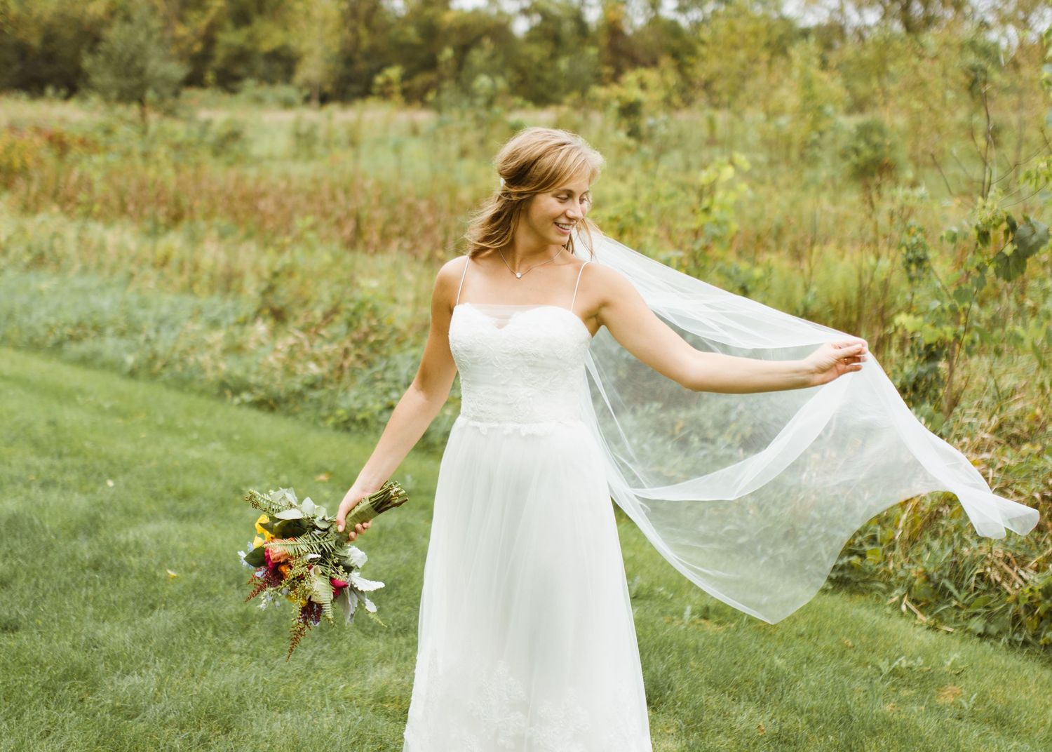 Bride in white gown twirling lace veil while holding colorful bouquet in green outdoor setting.