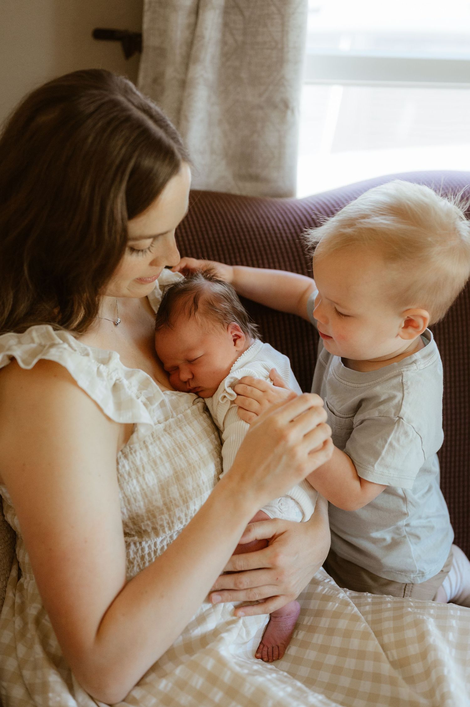 A mother sits in a chair holding her newborn baby while her young toddler son gently touches the infant's head.