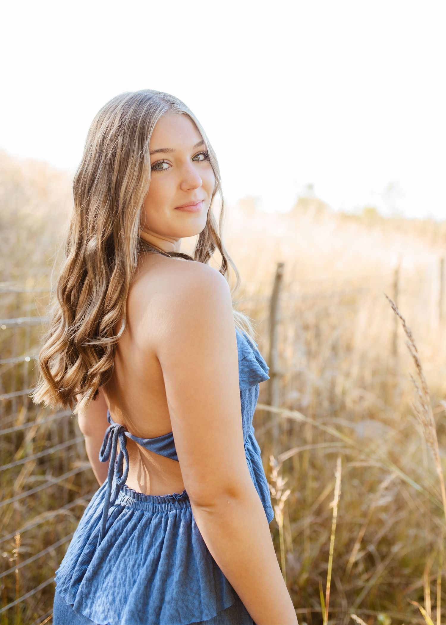 Young woman in a backless blue romper smiling over her shoulder in a golden grass field at sunset.