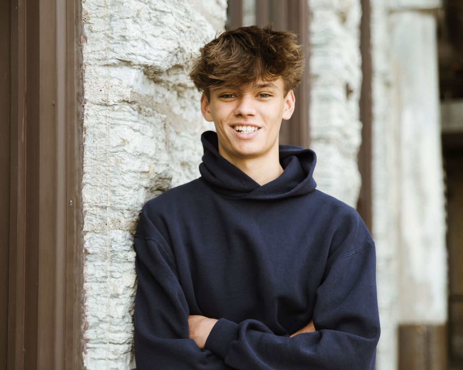 Smiling teen boy in navy hoodie leaning against a stone wall with arms crossed.