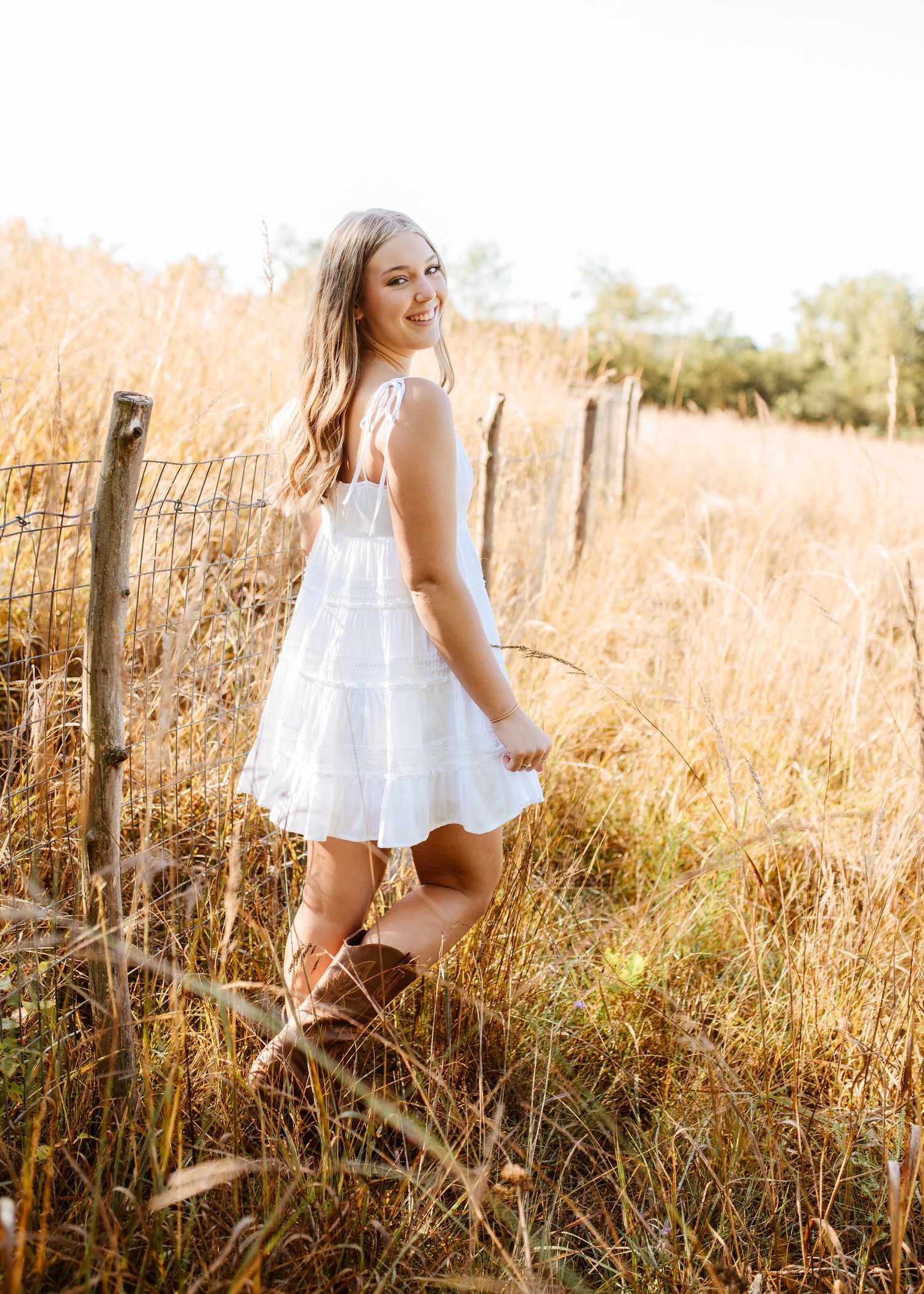 Young woman in a white mini dress and brown boots posing by a wooden fence in a golden field.