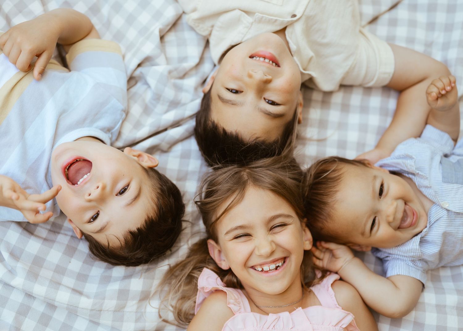 Four happy children lying in a circle on a blanket, laughing and smiling together in a joyful group photo.