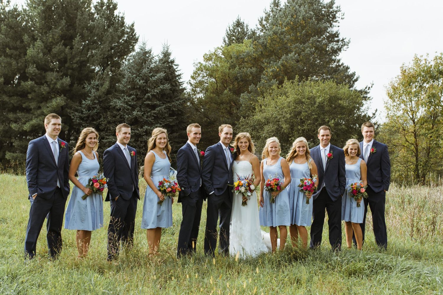 Wedding party posing in field with bridesmaids in light blue dresses and groomsmen in navy suits.