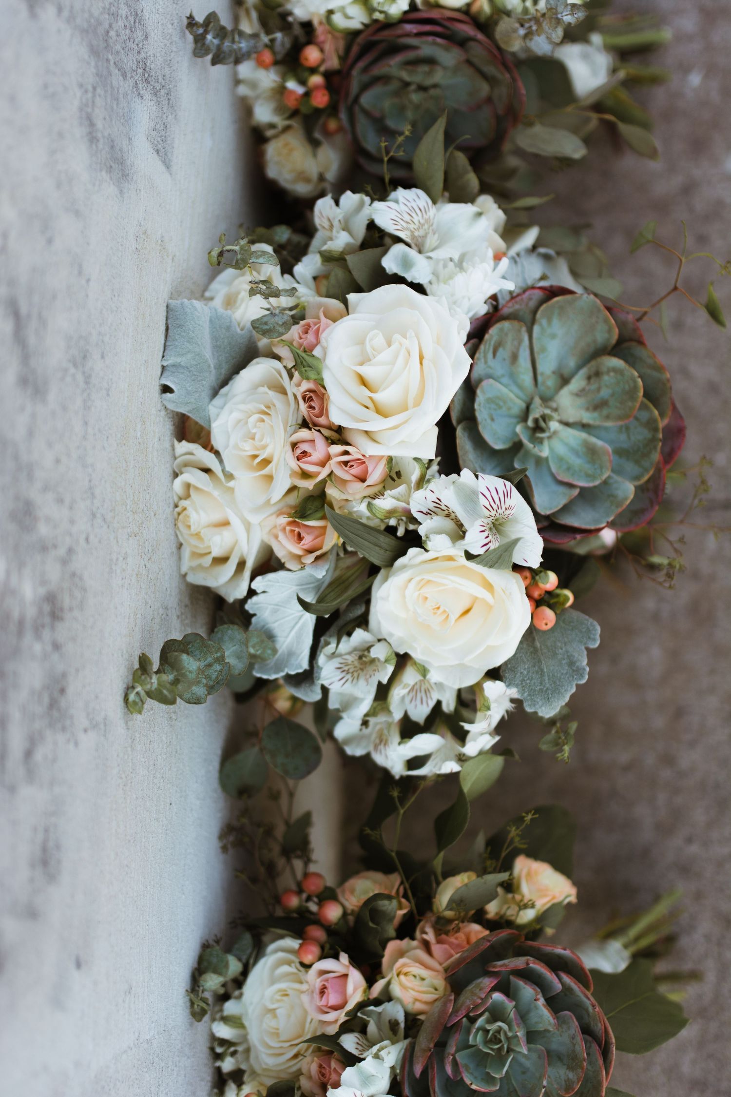 Romantic wedding bouquets featuring white roses, succulents, and eucalyptus resting against a wall.