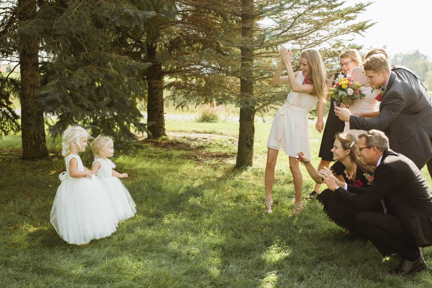 Wedding party poses outdoors with flower girls in white dresses on green grass surrounded by pine trees.