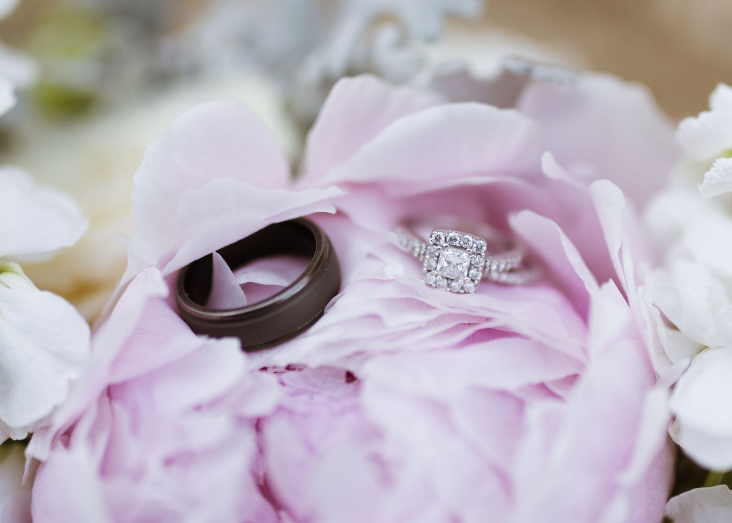 Diamond halo engagement ring and dark tungsten wedding band resting on a pink peony bloom.