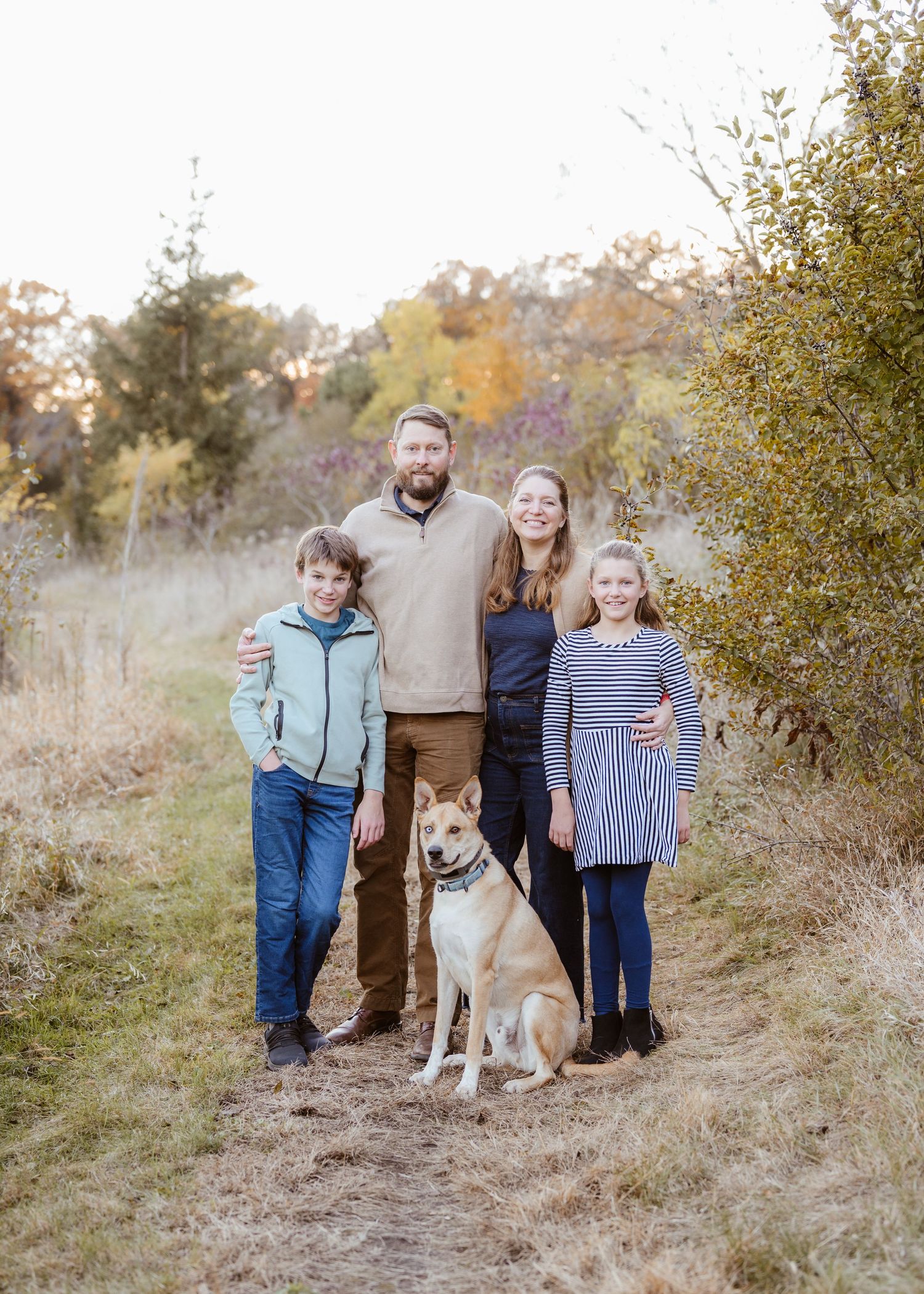 Happy family of four with two dogs posing on a nature trail surrounded by fall foliage.