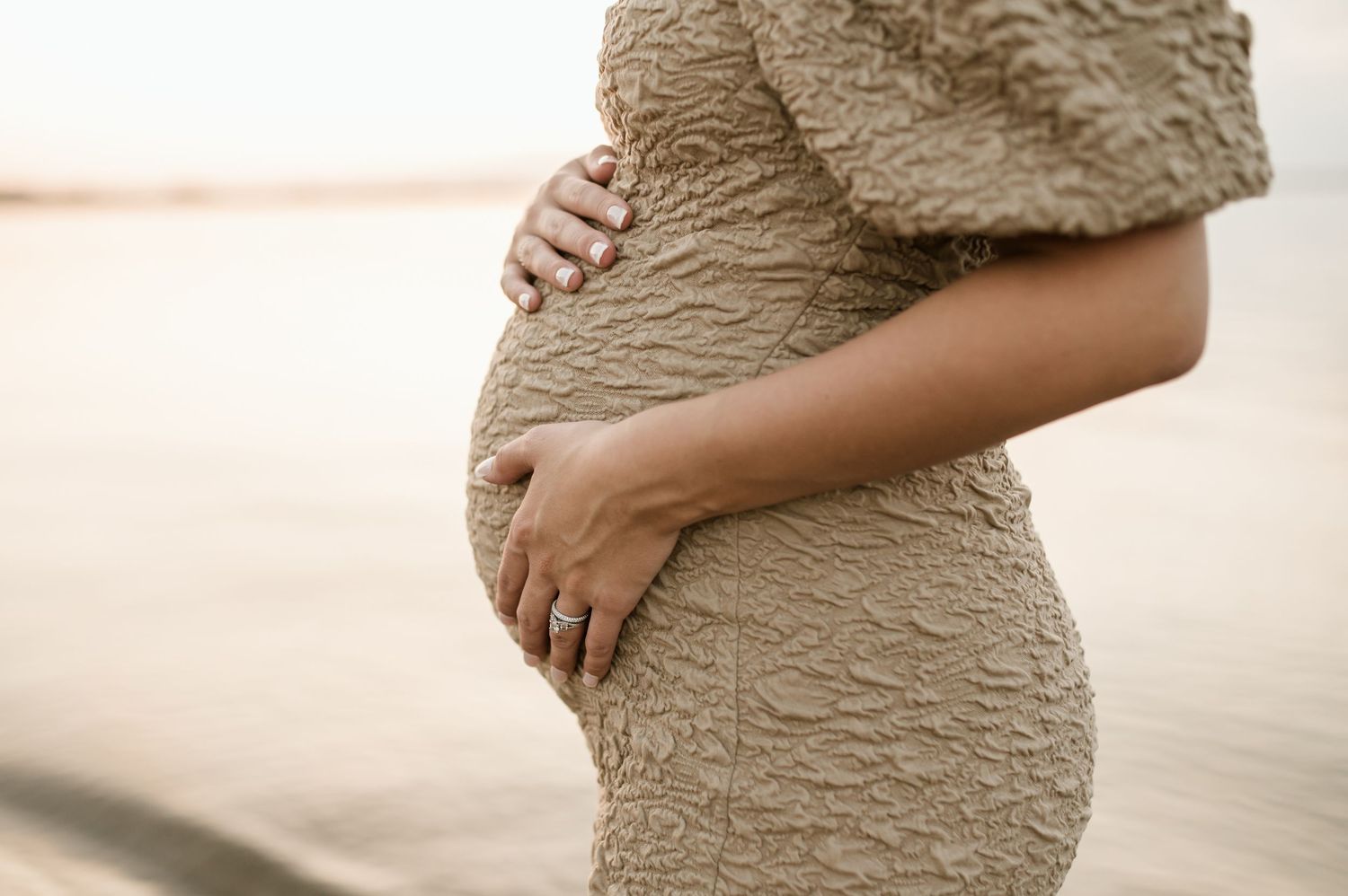 Close-up of a pregnant woman in a textured beige dress cradling her baby bump outdoors.