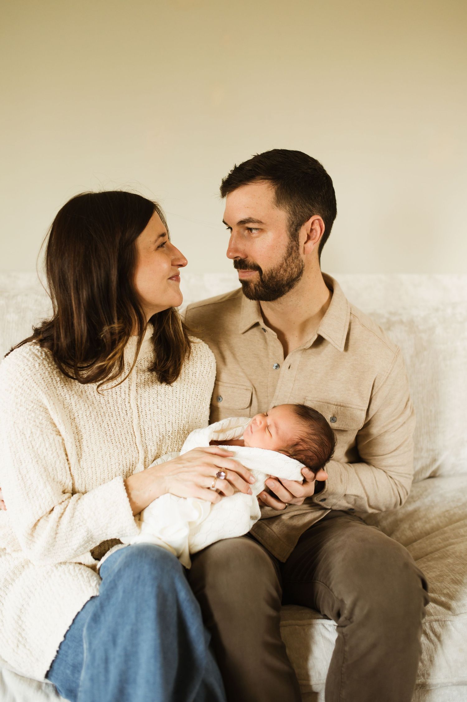 New parents sitting on couch smiling at each other while holding their swaddled newborn baby together.
