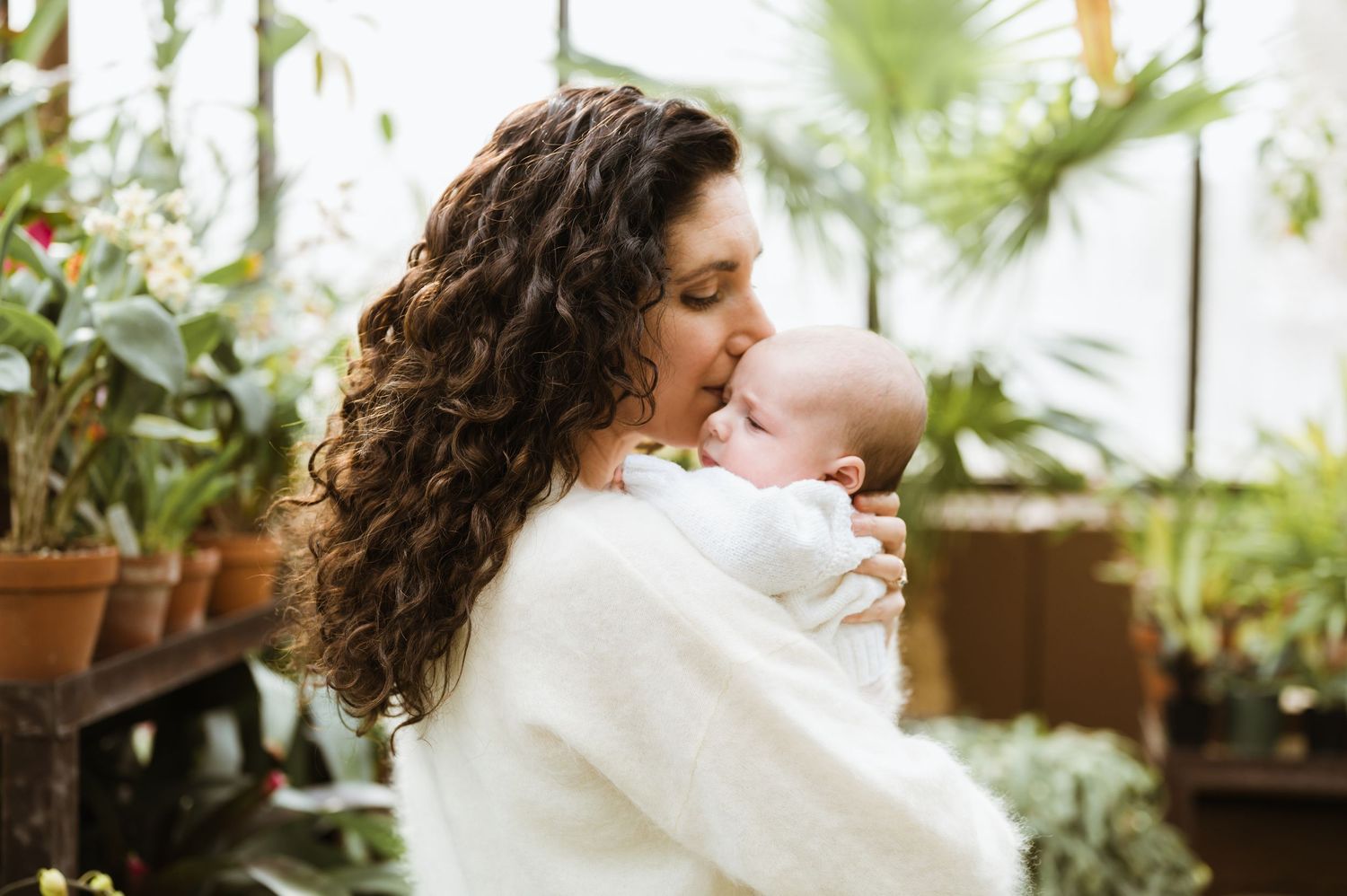 Curly-haired mother in white sweater kissing newborn baby in greenhouse.