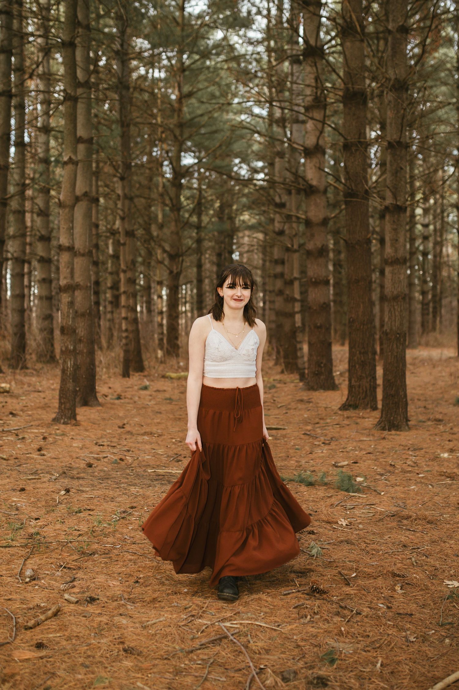 Young woman in white crop top and flowing rust maxi skirt posing in a pine forest.