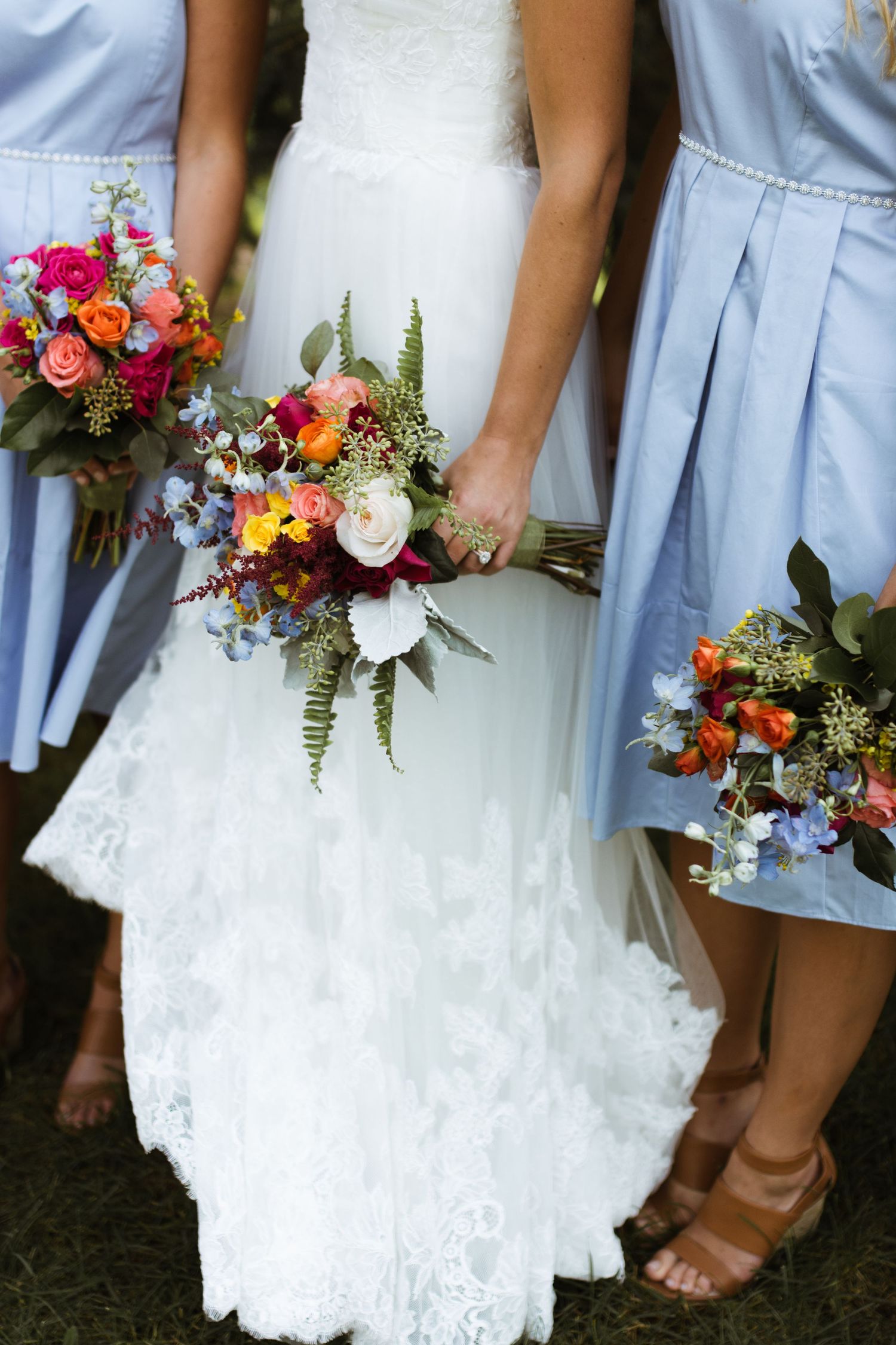 Bride and bridesmaids in light blue dresses holding vibrant colorful bouquets with ferns and wildflowers.