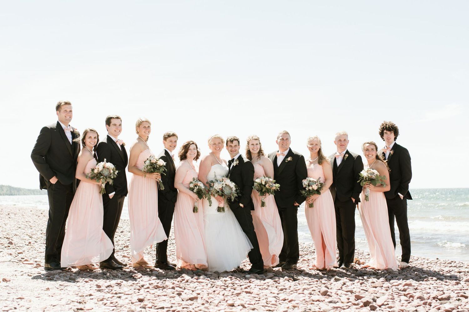 Wedding party in blush bridesmaid gowns and black suits posing together on a rocky beach.