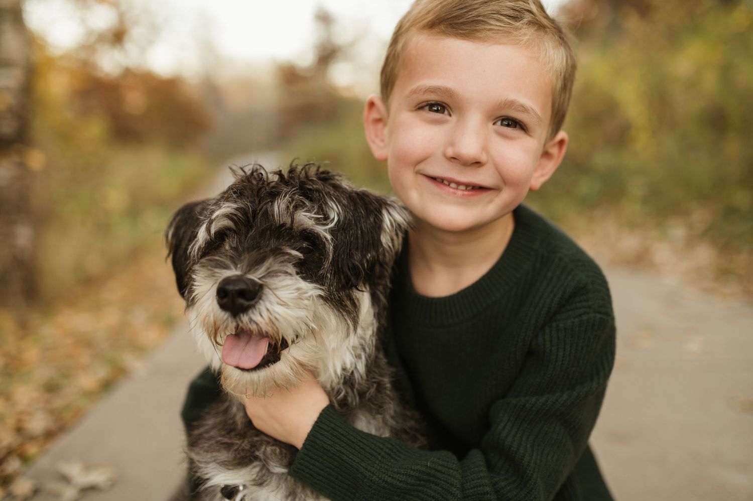 A smiling young boy hugs his fluffy black and white dog in an autumn outdoor setting.