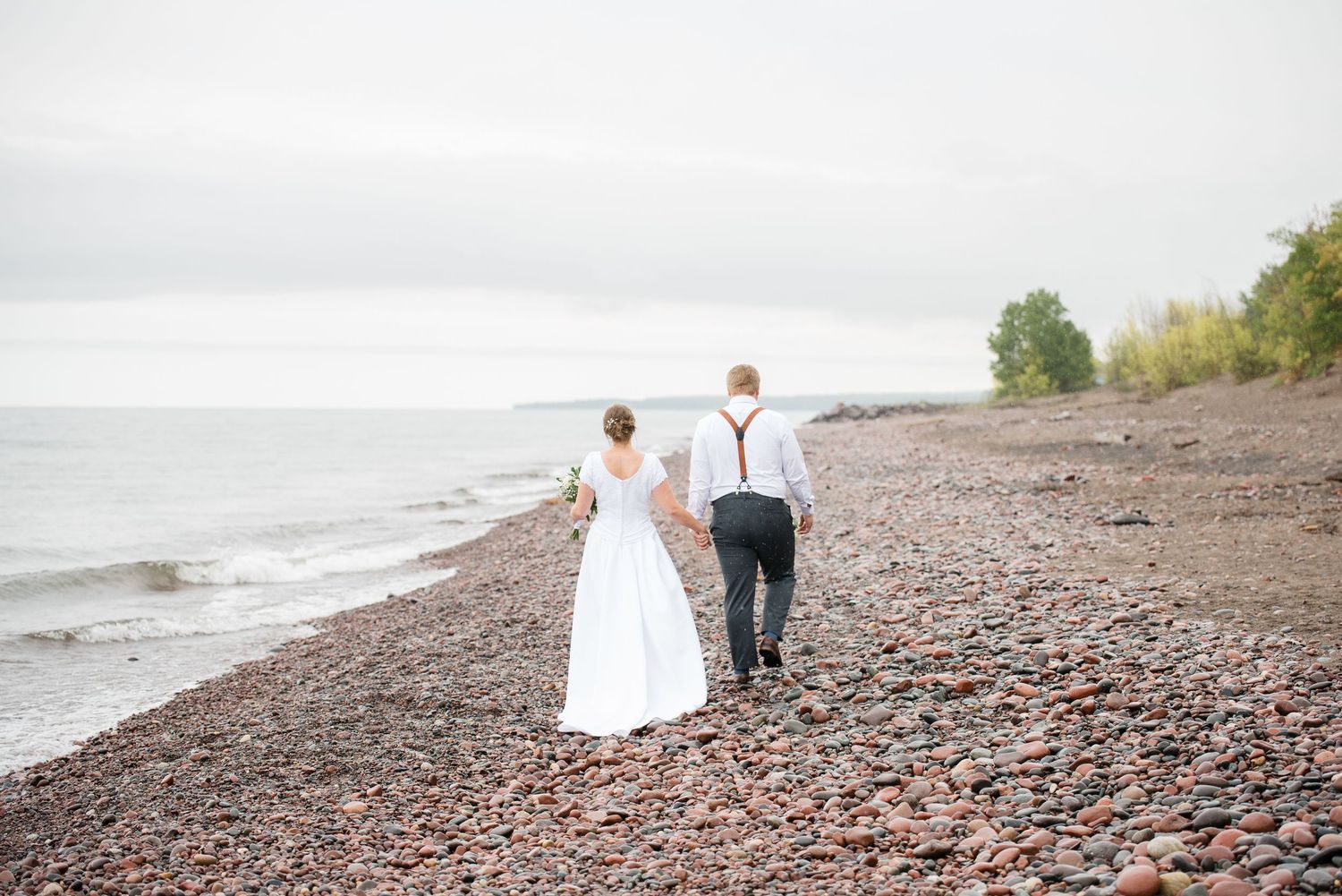 Bride and groom walk away holding hands along a rocky Lake Superior beach on their wedding day.