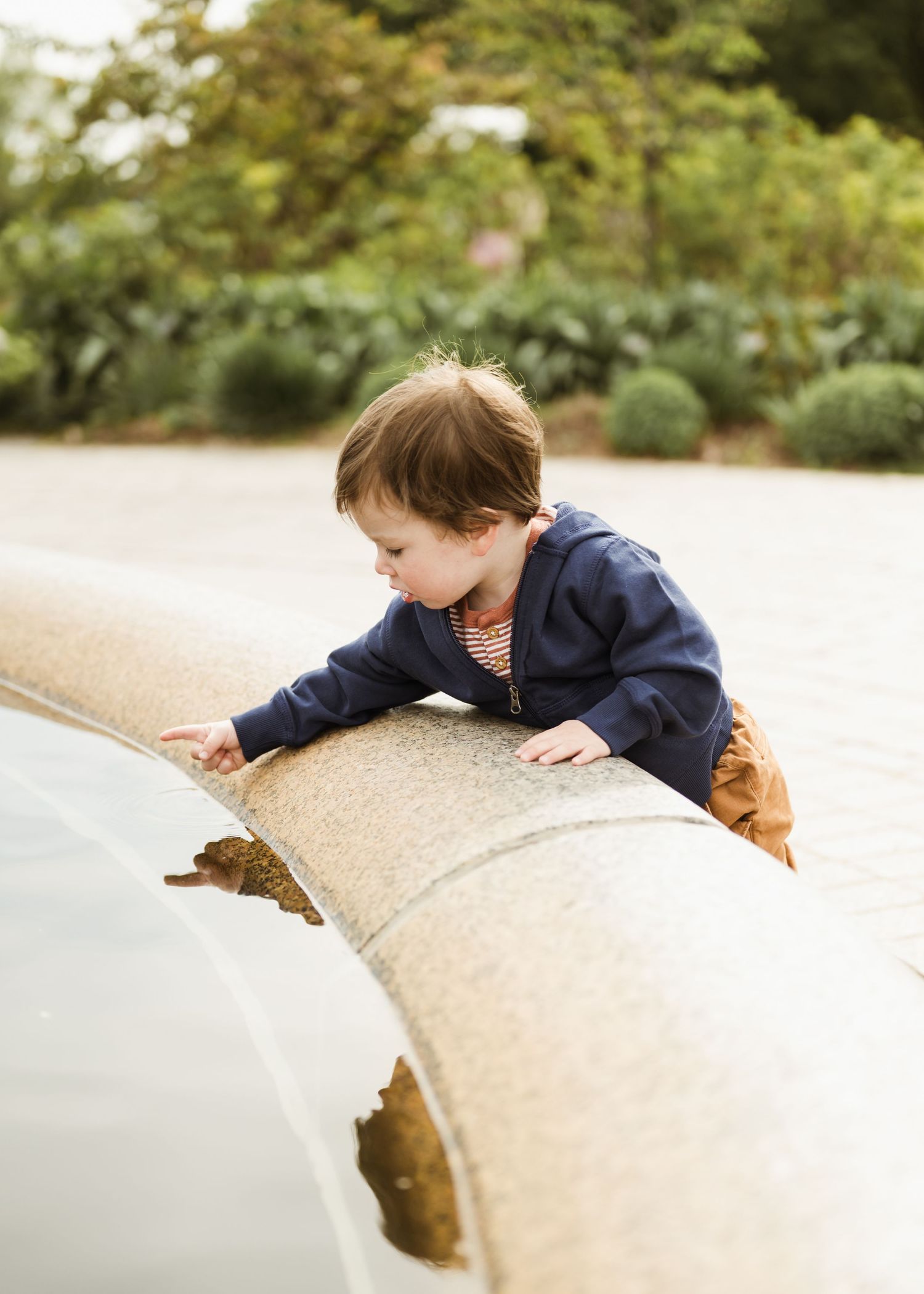 A young toddler in a blue hoodie leans over a stone fountain edge, exploring outdoors in a garden.