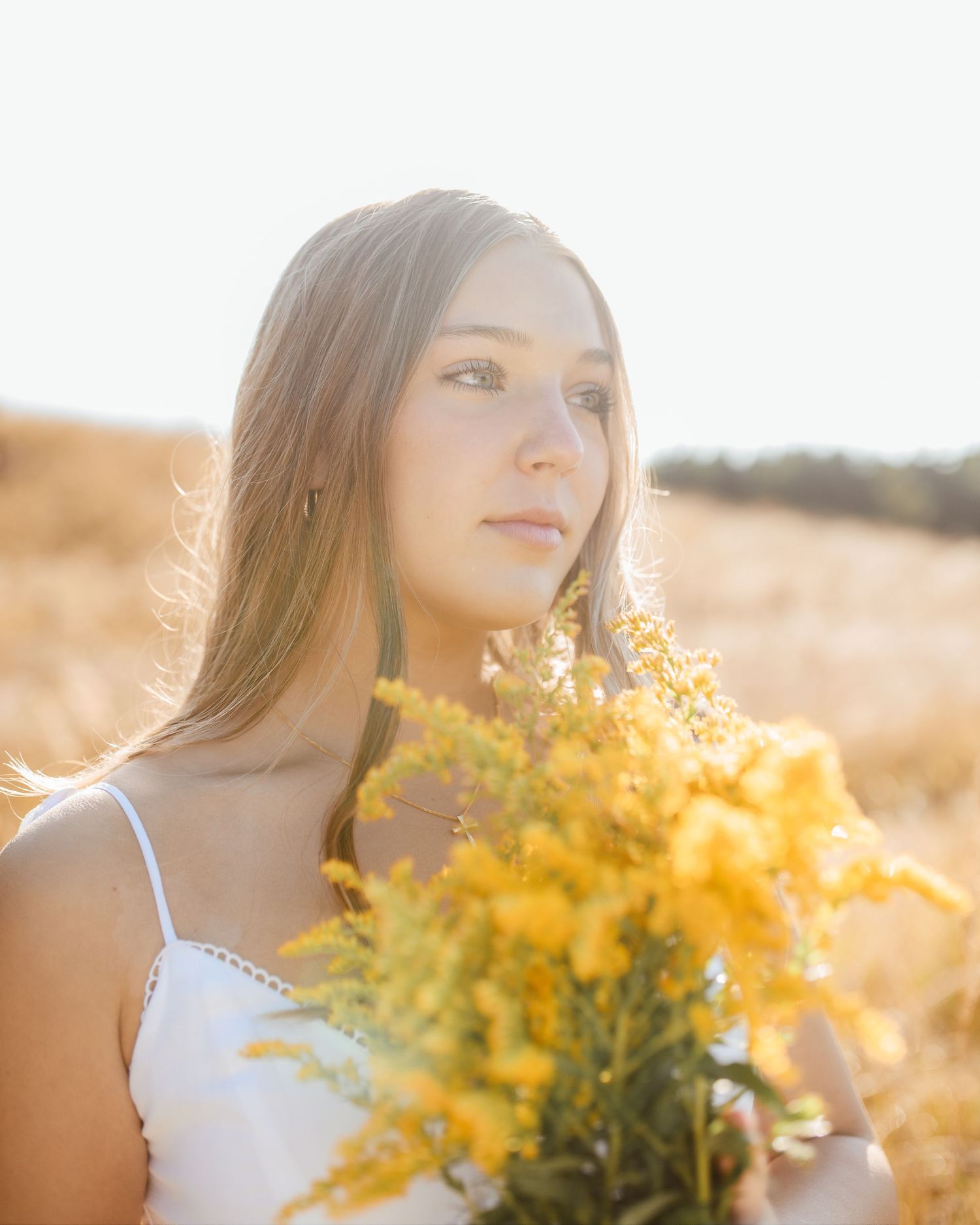 Young woman in a white dress holding yellow wildflowers in a sun-drenched golden field, gazing softly away.