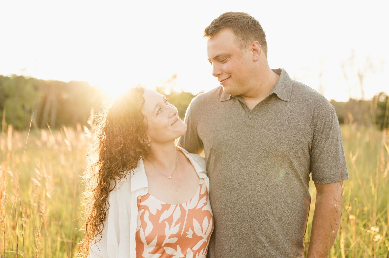 A smiling couple gazes at each other lovingly in a golden sunlit field during an engagement session.