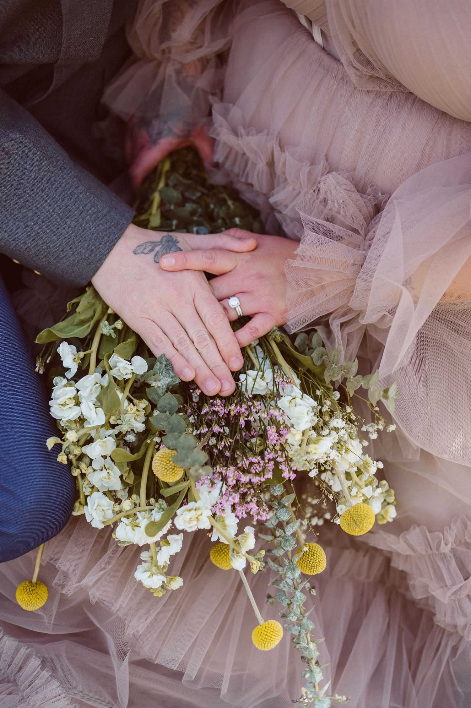 Close-up of bride and groom hands holding a wildflower bouquet with wedding ring visible.