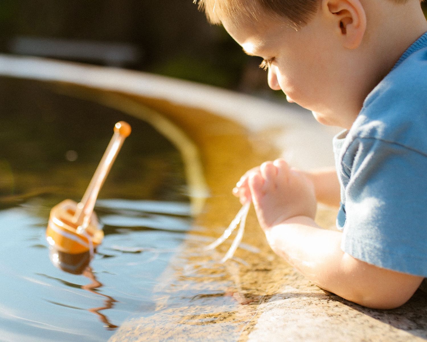 Young toddler boy in blue shirt playing with a small wooden toy sailboat floating in a sunlit outdoor water fountain.
