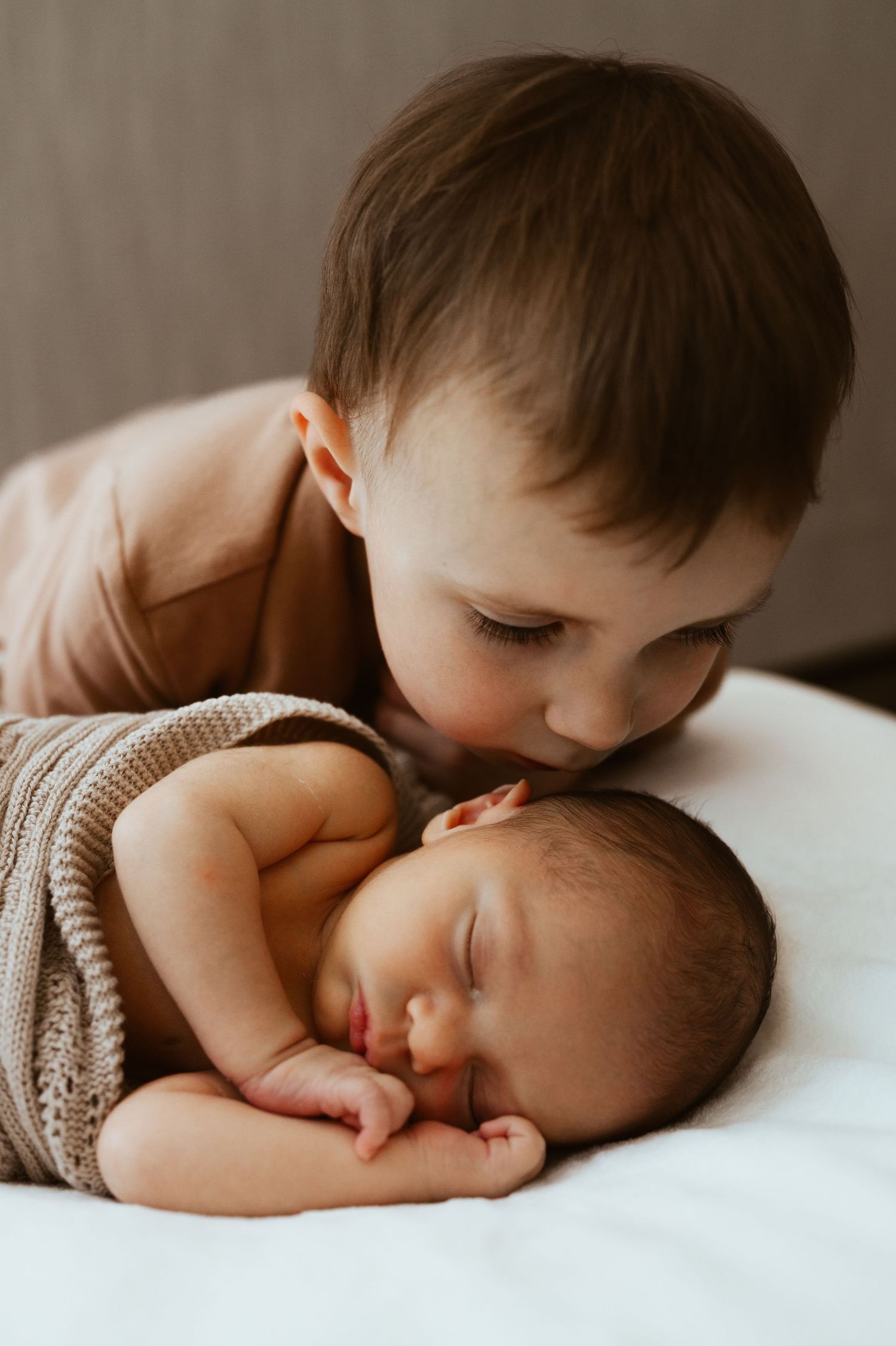 Toddler boy gently kissing sleeping newborn baby sibling wrapped in knit blanket on white surface.