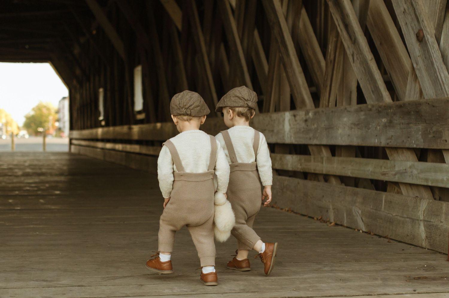 Two toddlers in matching outfits and caps walk together through a covered wooden bridge. Minnesota Family Photoshoot