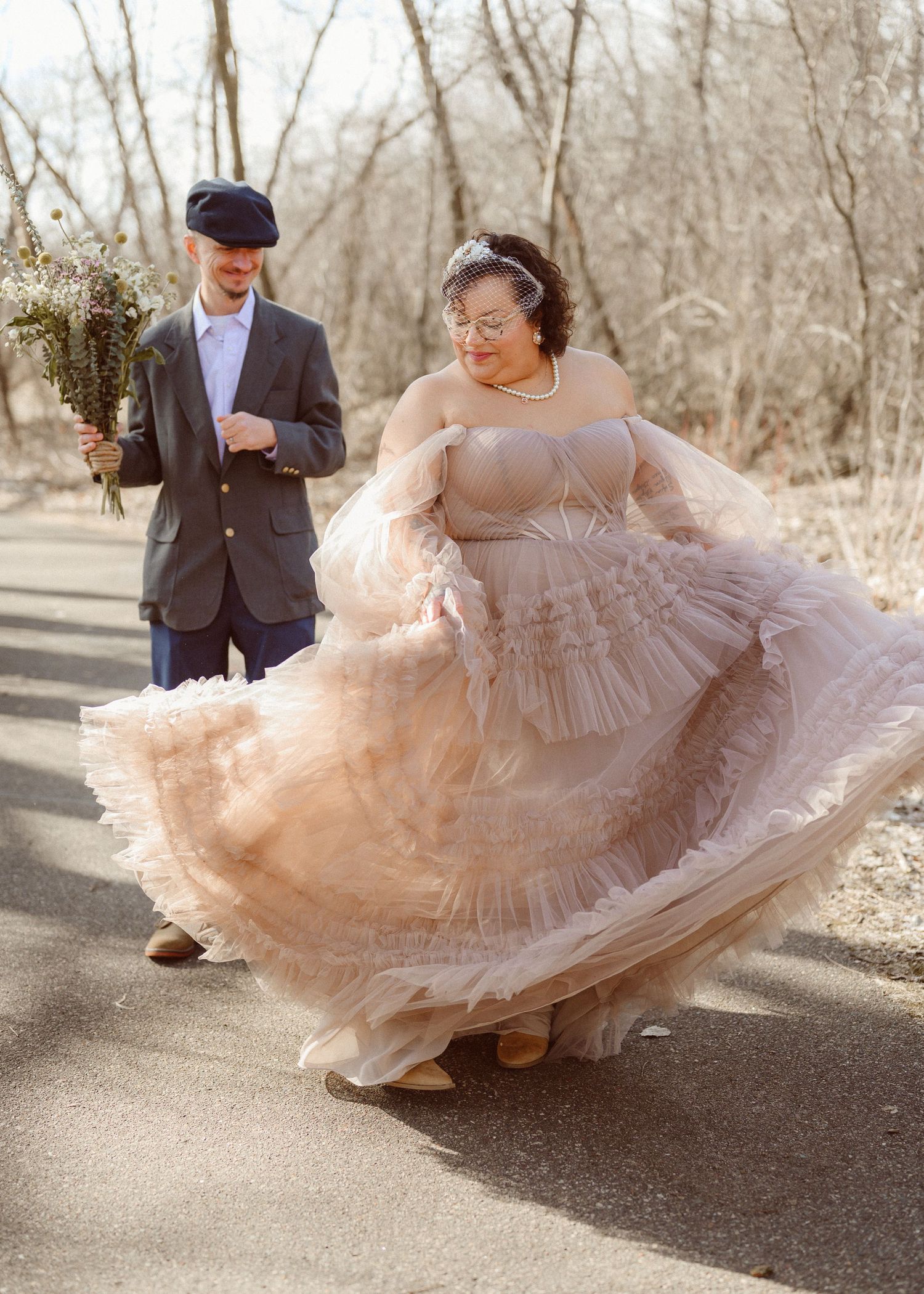Bride in blush ruffled tulle gown twirling on a path while groom watches holding a bouquet.