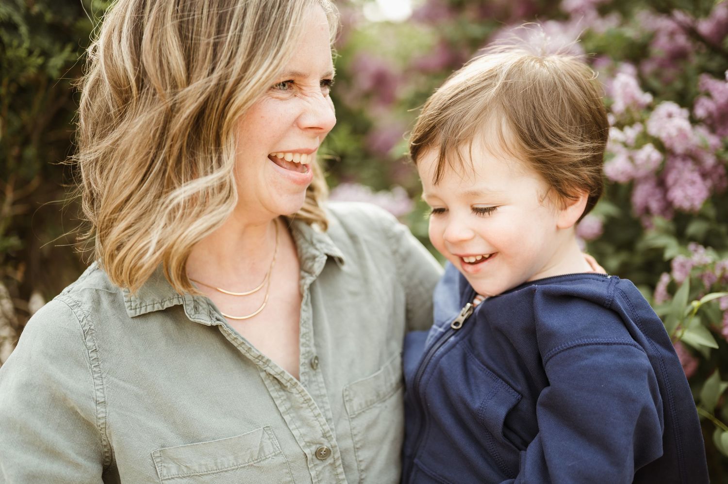 Smiling mother and toddler son sharing a joyful moment outdoors with blooming pink flowers behind them.